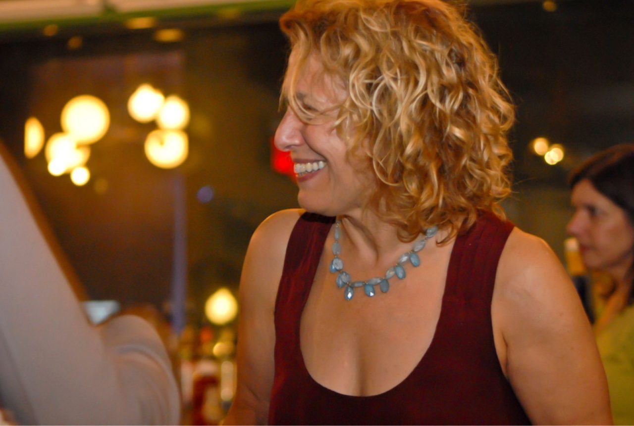 Smiling woman with curly hair and a necklace in a warmly lit setting.